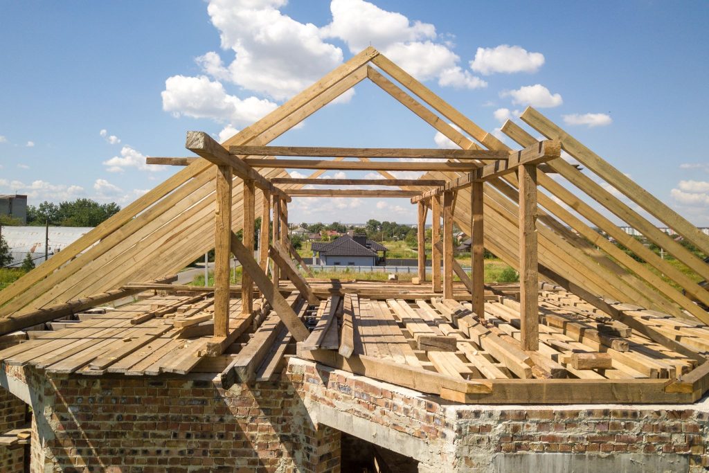 A roof is stripped back to supporting beams, clearly a construction zone. The sky is blue and mostly clear, showing off gorgeous clear summer weather. -- roof replacement summer